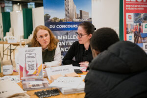 Deux femmes sont assises à une table lors d’un événement professionnel, discutant avec un homme. Des brochures et une pancarte indiquant « Kaufman & Broad » sont visibles. L’atmosphère est professionnelle.